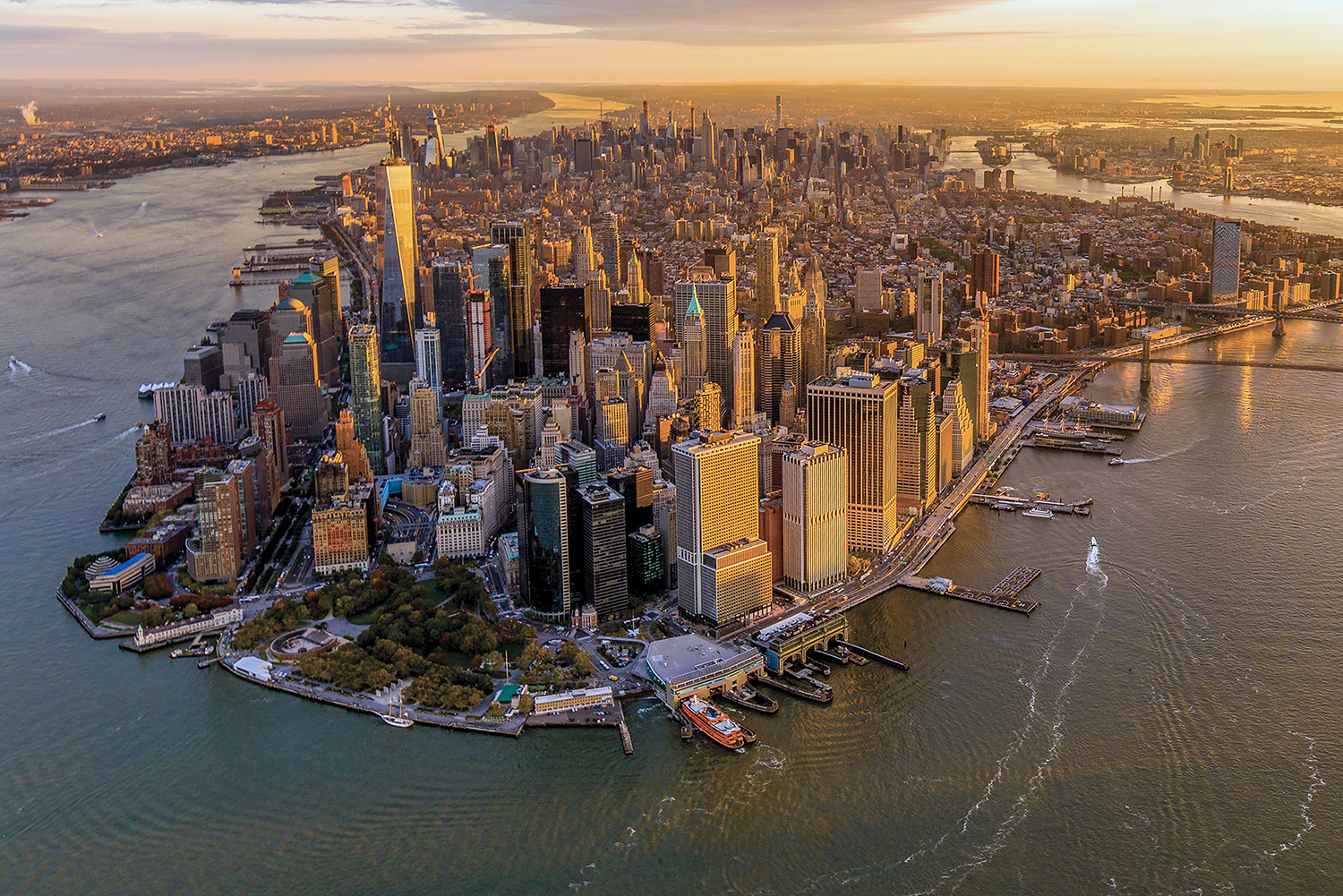Aerial view of Lower Manhattan at sunset, showing densely packed skyscrapers, surrounding rivers, docks, and boats, with the rest of New York City extending into the distance.