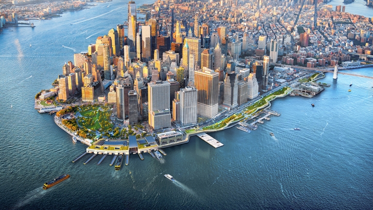 Aerial view of Lower Manhattan, New York City, with tall skyscrapers, waterfront parks, boat traffic, and surrounding rivers visible under a partly cloudy sky.