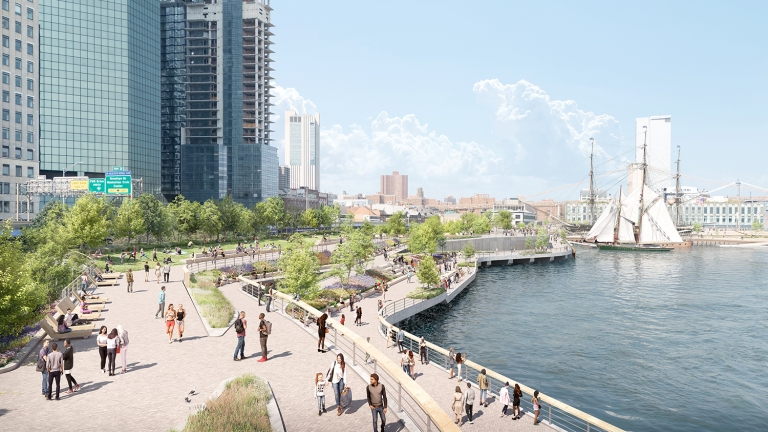 A waterfront park with walking paths, people strolling and relaxing, trees, and city skyscrapers in the background. A historic sailing ship is docked nearby on the river under a partly cloudy sky.