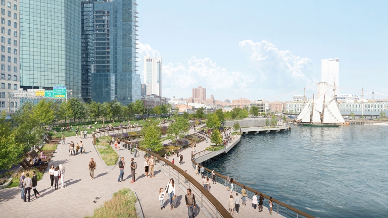 People walk along a waterfront park with green spaces, trees, and benches. Tall modern buildings line the left side, while a historic sailing ship and bridges are visible on the river to the right under a bright sky.