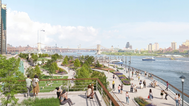 People walk, relax, and sit along a landscaped riverside park with trees and flowers, overlooking a wide river with boats and a distant bridge under a bright, partly cloudy sky.