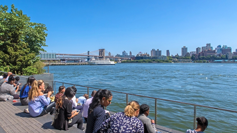 People sit on steps by the waterfront on a sunny day, looking out at the river, boats, and the New York City skyline, including the Brooklyn Bridge.