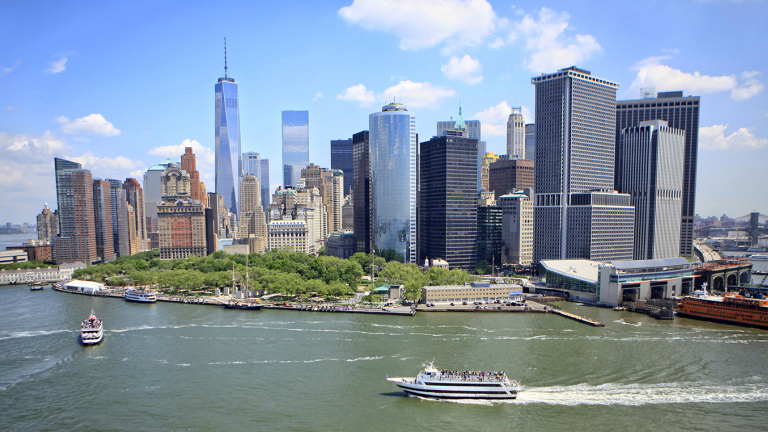 Aerial view of Lower Manhattan, New York City, showing skyscrapers, ferries on the Hudson River, and green spaces under a bright blue sky with scattered clouds.