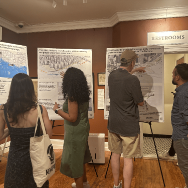 Five people stand in a small gallery, closely examining and discussing large informational posters about urban planning and flood resilience displayed on easels; a “Restrooms” sign is visible on the back wall.