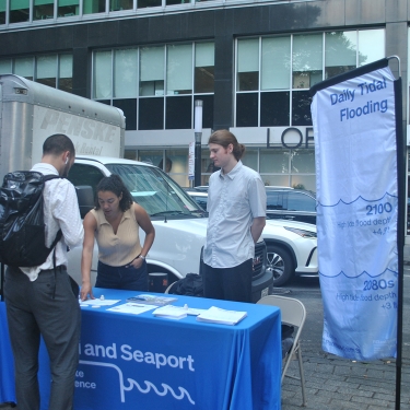 Three people at an outdoor information table about daily tidal flooding, with banners and brochures. The table is on a city sidewalk, with buildings, a truck, and a Starbucks in the background.