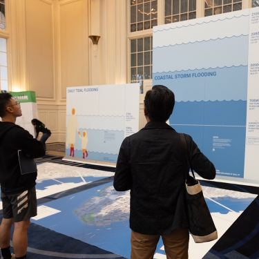 Two people stand indoors facing large informational displays about flooding, with graphics and text illustrating daily tidal and coastal storm flooding projections. Other attendees are visible in the background.