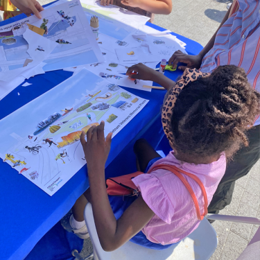 A young girl sits at a blue table working on a colorful drawing with others. Art supplies and illustrated papers are spread out, with children’s hands visible as they participate in the creative activity outdoors.