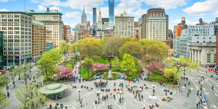 Aerial view of New York&#039;s Union Square, a lively urban park surrounded by tall buildings under a blue sky with clouds. The park features lush green trees, blooming pink flowers, a central fountain, and numerous people walking and sitting.
