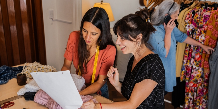 Two women fashion designers are looking at a sheet of paper and fabric samples in a sewing room. Another woman in the background is talking on the phone by a clothing rack.