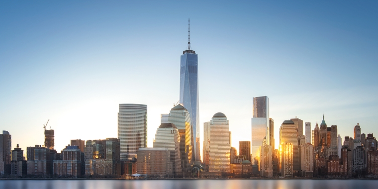 Skyline of New York City with modern high-rise buildings reflecting in calm water, under a clear blue sky at sunset.