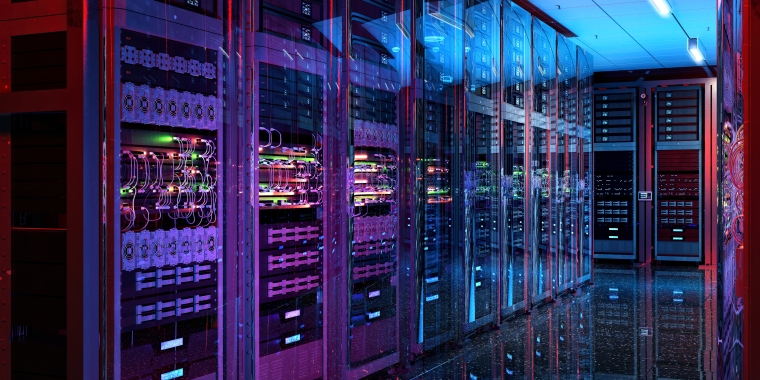 A modern server room with multiple server racks on either side. The racks are filled with cables and illuminated by colorful LED lights, casting a vibrant glow on the reflective floor. The ceiling has bright overhead lights.