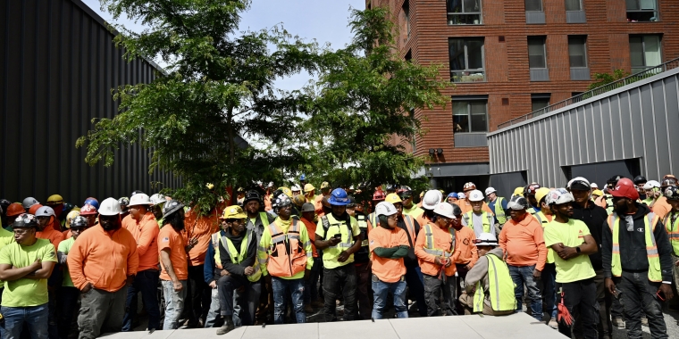 Large group of construction workers wearing safety vests and hard hats stand together outside Hunts Point Peninsula, some with arms crossed, listening attentively. A leafy tree is in the background.