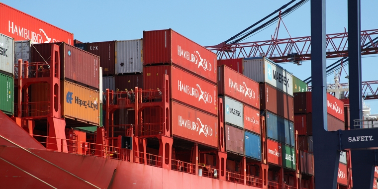 A large red cargo ship loaded with colorful shipping containers is docked at a port, with cranes and blue sky visible in the background.