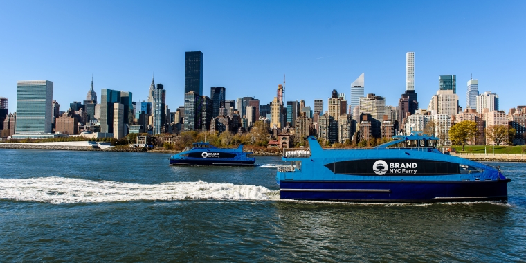 Two blue NYC ferries, each labeled BRAND NYC FERRY, travel on the river. The New York City skyline, including tall skyscrapers and clear blue sky, is in the background.