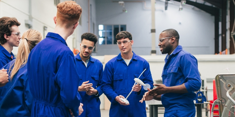 Group of men wearing blue coveralls and safety glasses stand together in a workshop, listening to a man who appears to be explaining something while holding a tool. Industrial equipment is visible in the background.