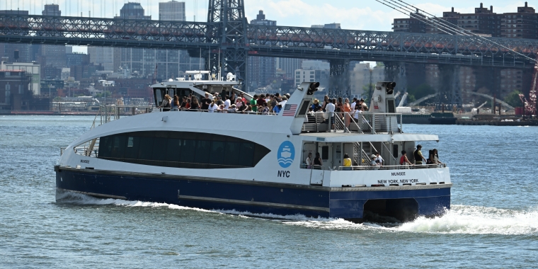 A white and blue NYC ferry with passengers on board travels along the water, with a New York City skyline, buildings, and a bridge visible in the background. Photo courtesy of John Vital.