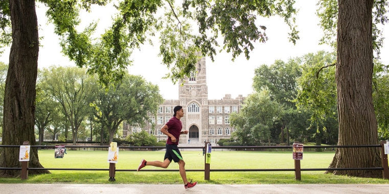 A man in athletic wear jogs on the Fordham University campus along a path lined with trees, with a large, historic stone building and green lawn in the background on a bright, clear day.
