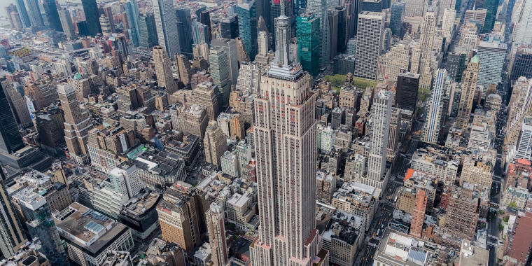 Aerial view of the Empire State Building surrounded by densely packed skyscrapers and New York City streets in Midtown Manhattan on a clear day.