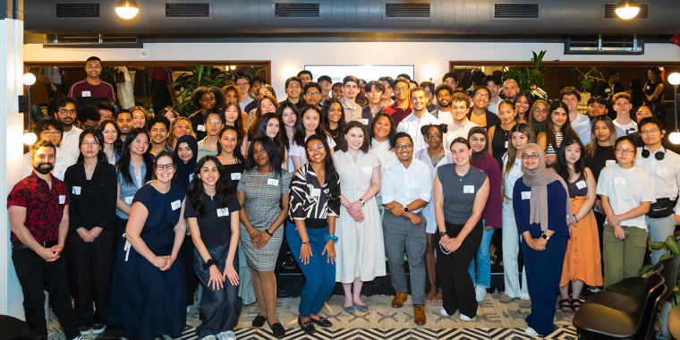 A large, diverse group of people poses together and smiles for a group photo in a brightly lit, modern indoor space with patterned flooring and plants in the background.
