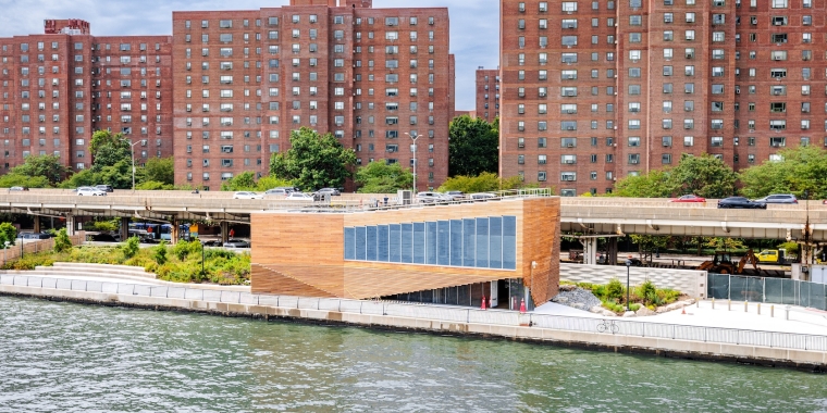 A modern wooden building for Solar One Environmental Education Center sits by the water with greenery around it, while tall red-brick apartment buildings and a highway with cars are visible in the background under a partly cloudy sky.