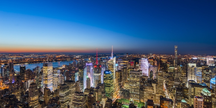 Aerial view of a brightly lit city skyline at dusk, with tall skyscrapers, illuminated buildings, and a river visible in the distance under a deep blue sky.