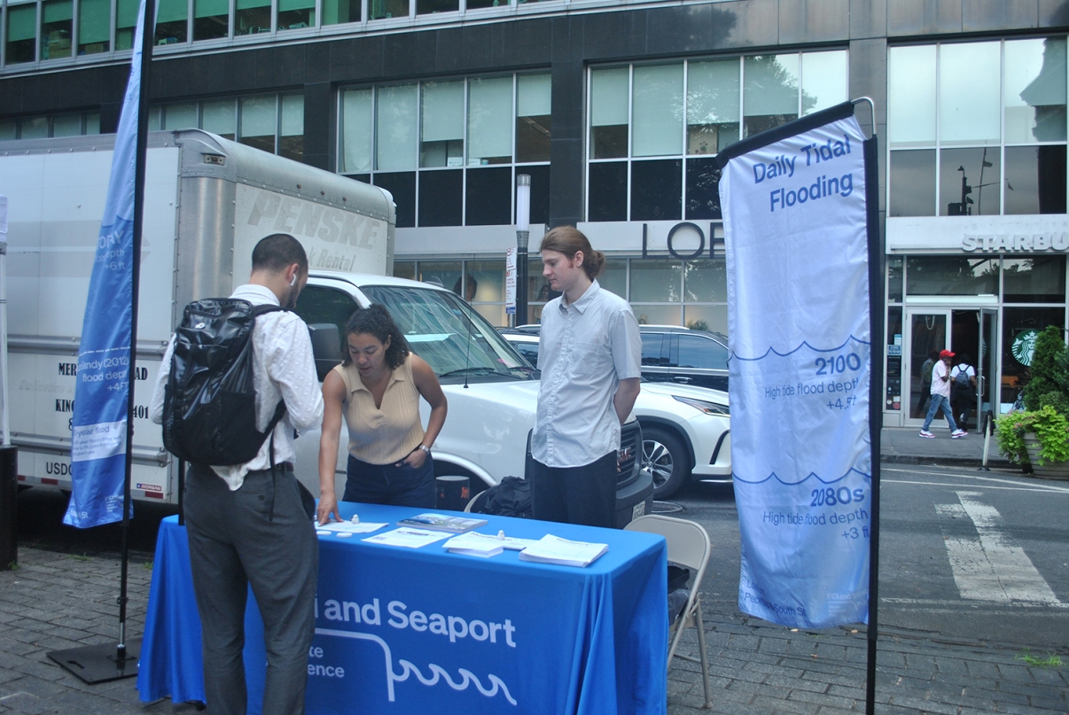 Three people at an outdoor information table about daily tidal flooding, with banners and brochures. The table is on a city sidewalk, with buildings, a truck, and a Starbucks in the background.