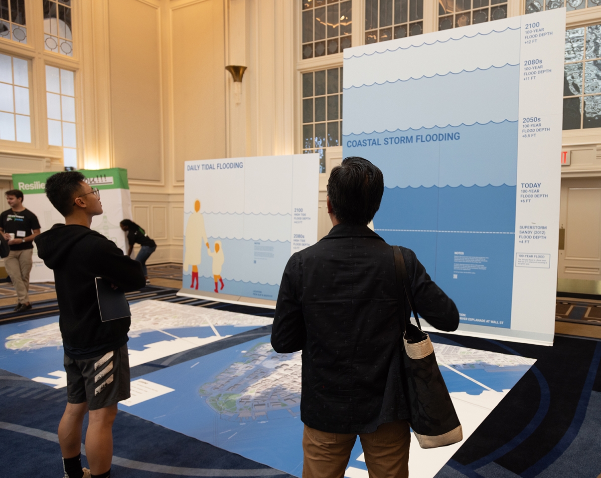 Two people stand indoors facing large informational displays about flooding, with graphics and text illustrating daily tidal and coastal storm flooding projections. Other attendees are visible in the background.