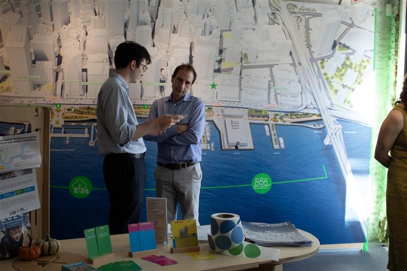 Two men stand talking in front of a large, detailed city and harbor map. A table with colorful brochures and models is in the foreground. A third person, partially visible, stands to the right.