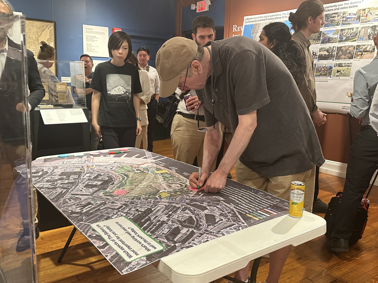 A man leans over a table with a large map, writing or drawing on it with a red marker. Several people observe, and informational posters are displayed in the background. A can of drink sits on the table.